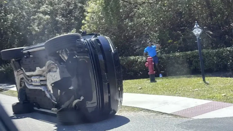 An overturned Land Rover on a Florida roadside with emergency lights in the background.